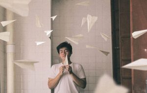 Boy in white shirt letting go of a paper airplane while standing in the center of a white wall with his eyes closed and other paper airplanes fly around